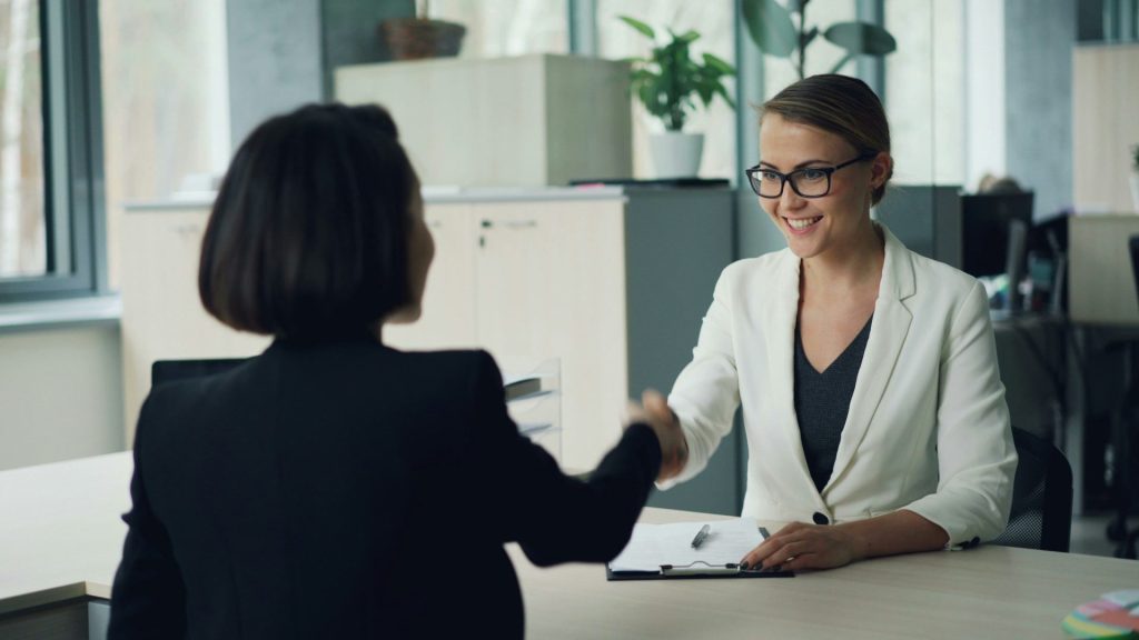 Zwei Frauen, die sich über einen Schreibtisch hinweg die Hände schütteln